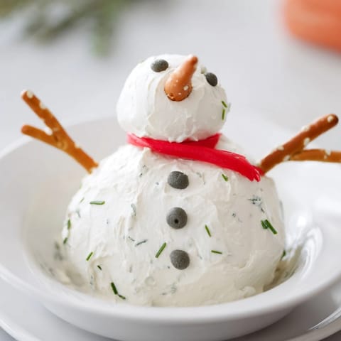 A close-up of a festive Melted Snowman Dip, complete with a carrot nose and olive eyes.