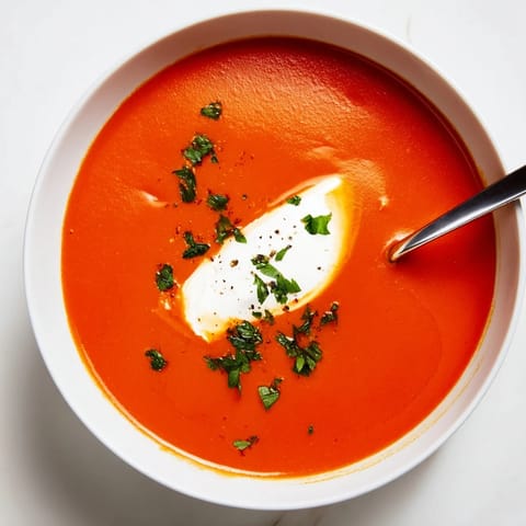 Spoon dipping into creamy Roasted Red Pepper Soup, revealing silky texture, with crusty bread and olive oil nearby.