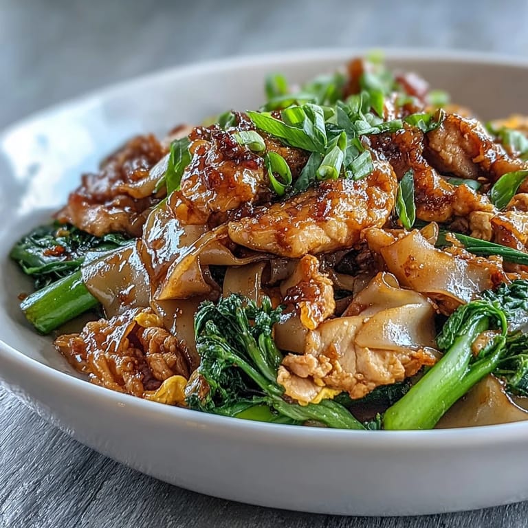 A close-up of Authentic Thai Pad See Ew, showcasing glossy noodles, chicken, and Chinese broccoli on a plate. 